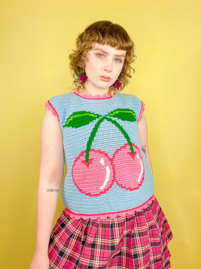 A woman posing wearing a crocheted vest in baby blue with a pink cherry on the front, paired with a pink kilt skirt.