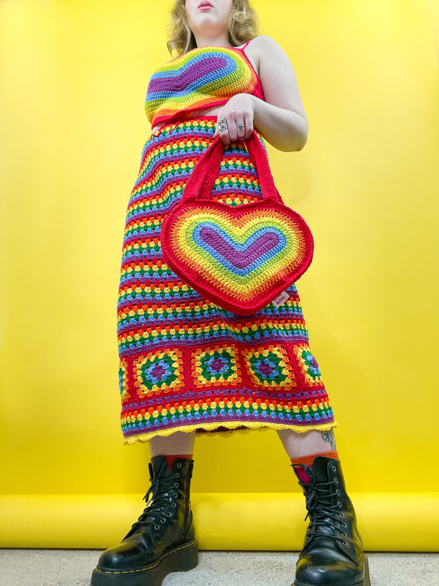 Low view of a woman wearing a matching rainbow top and skirt set paired with a heart shaped rainbow crochet bag.