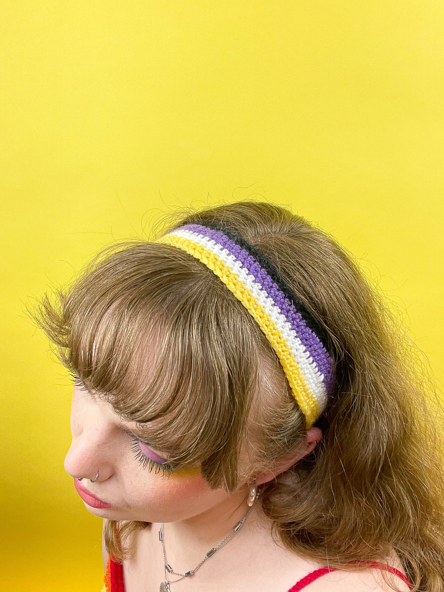 Above view of a woman wearing a headband with the non binary flag colours (yellow, white, purple and black) in front of a yellow background.