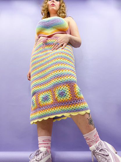 Below view of a woman wearing a crochet midi skirt in pastel rainbow colours and a matching heart shaped top paired with lilac platform shoes.