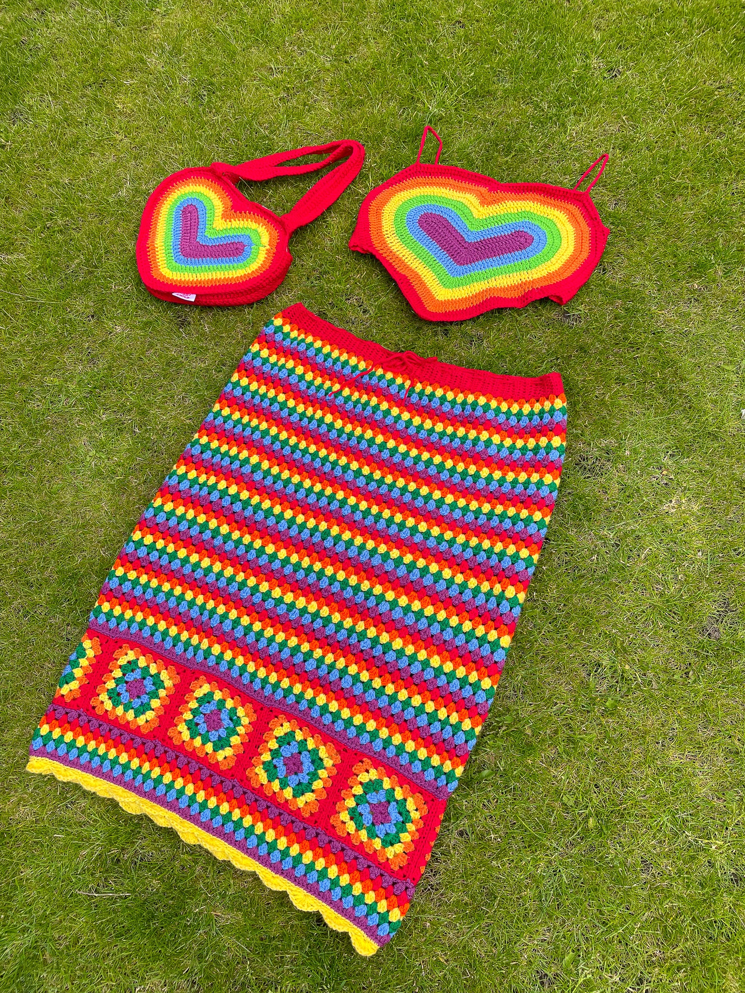 A crocheted rainbow set lying together on the grass, with a heart shaped crop top with straps, a striped granny square skirt and a heart shaped shoulder bag.