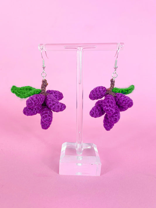 A pair of crocheted earrings shaped like grape vines in purple with a green leaf attached to them hung up on an earring stand in front of a pink background.