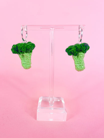 Pair of silver hoop earrings with crocheted mini broccolis attached to them hanging on a clear earring stand.