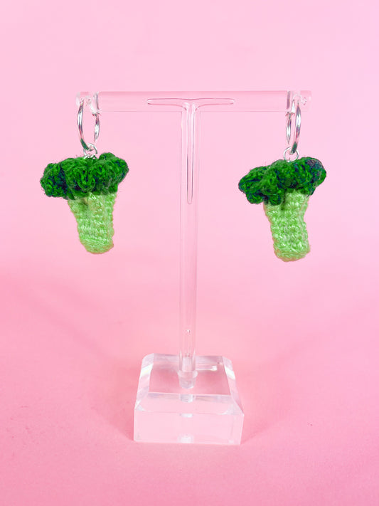 Pair of silver hoop earrings with crocheted mini broccolis attached to them hanging on a clear earring stand.