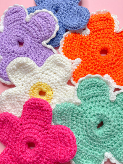 Six crochet flower scrunchies in different colours laying on top of each other on a pink background.