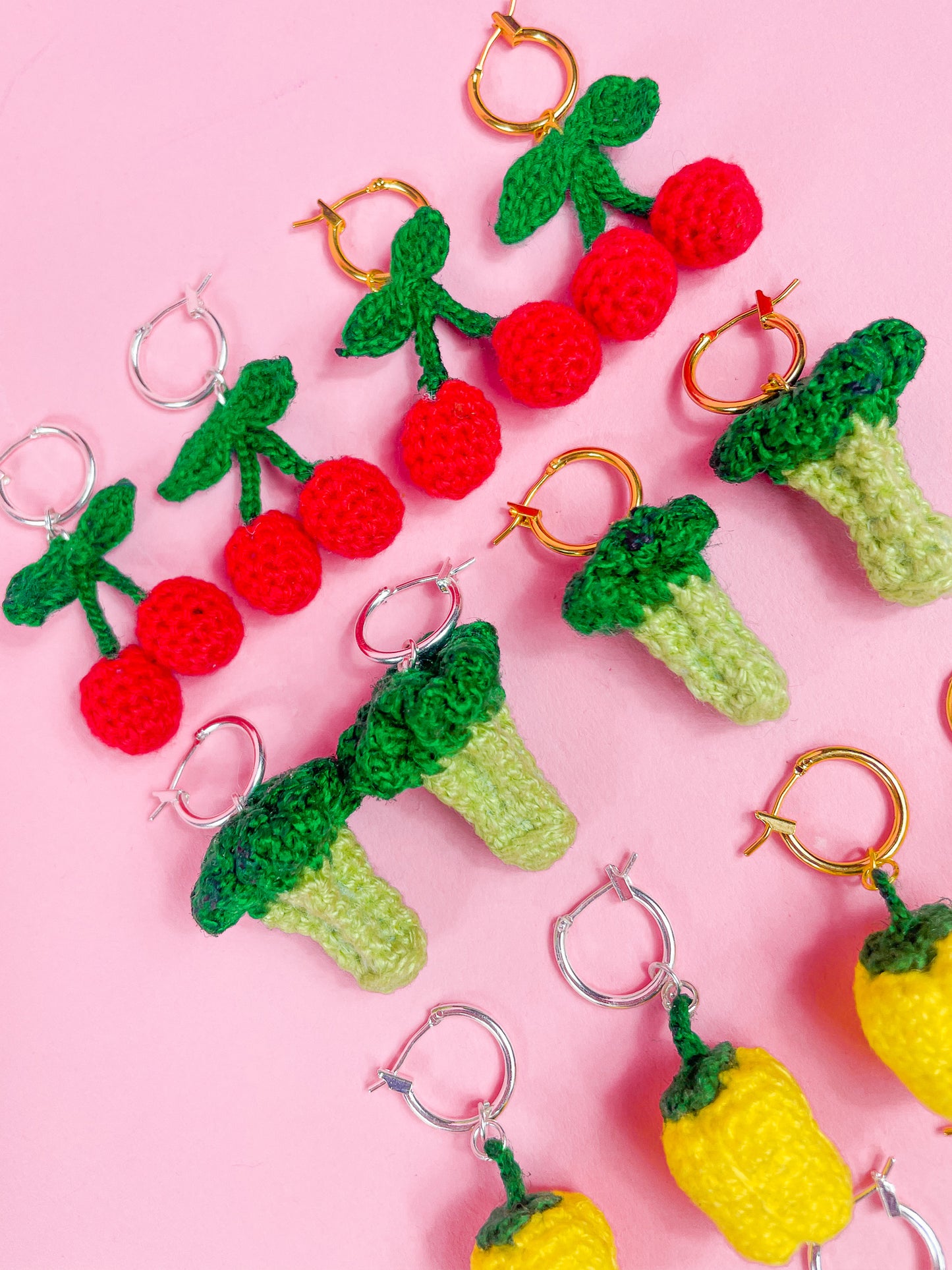 Six pairs of earrings in various fruit and vegetable shapes (cherries, broccolis and bell peppers), lying together in rows.