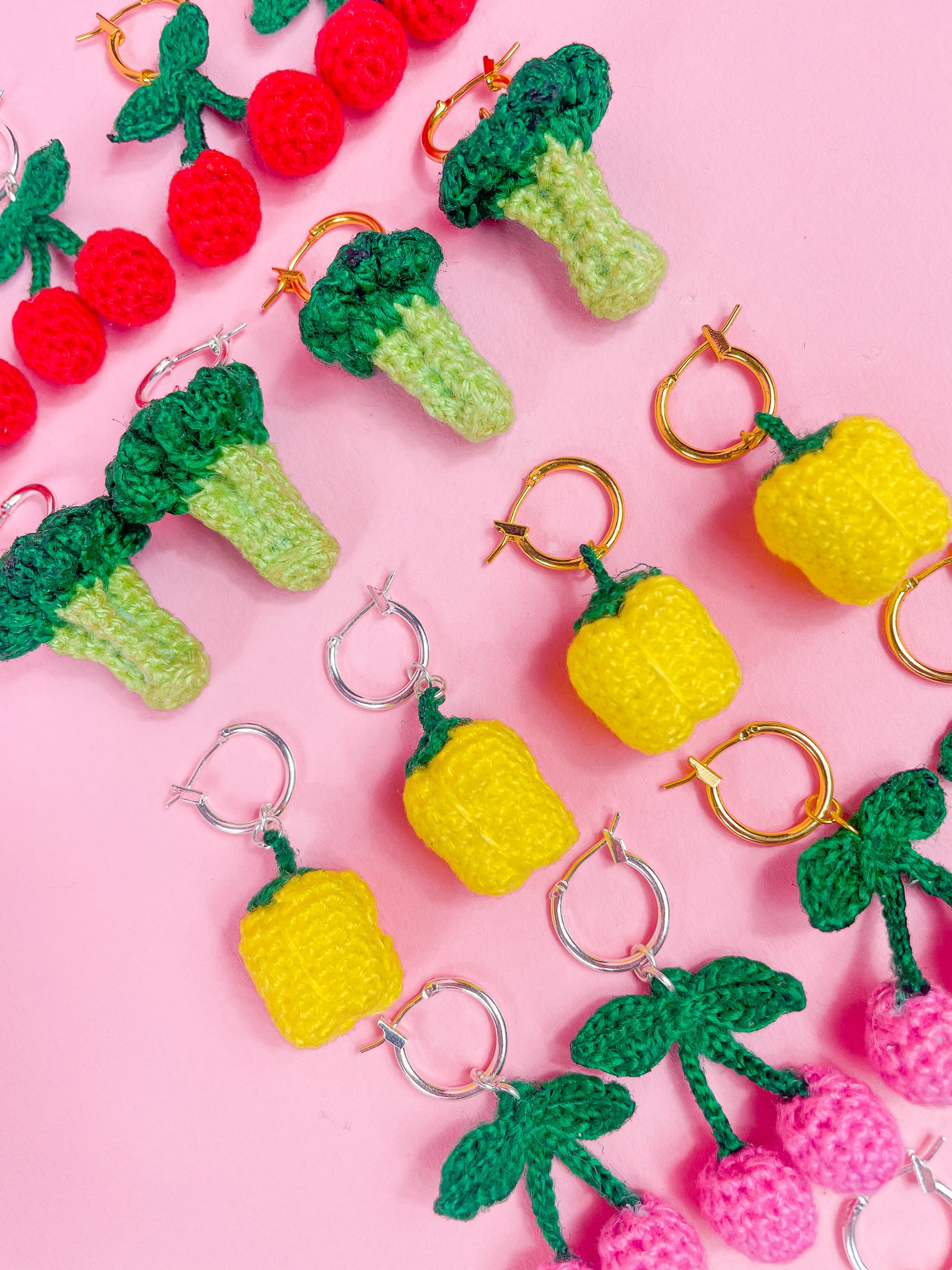 Eight pairs of crocheted earrings all shaped like different fruits (cherries, broccolis and bell peppers), lying together in rows on a pink background.