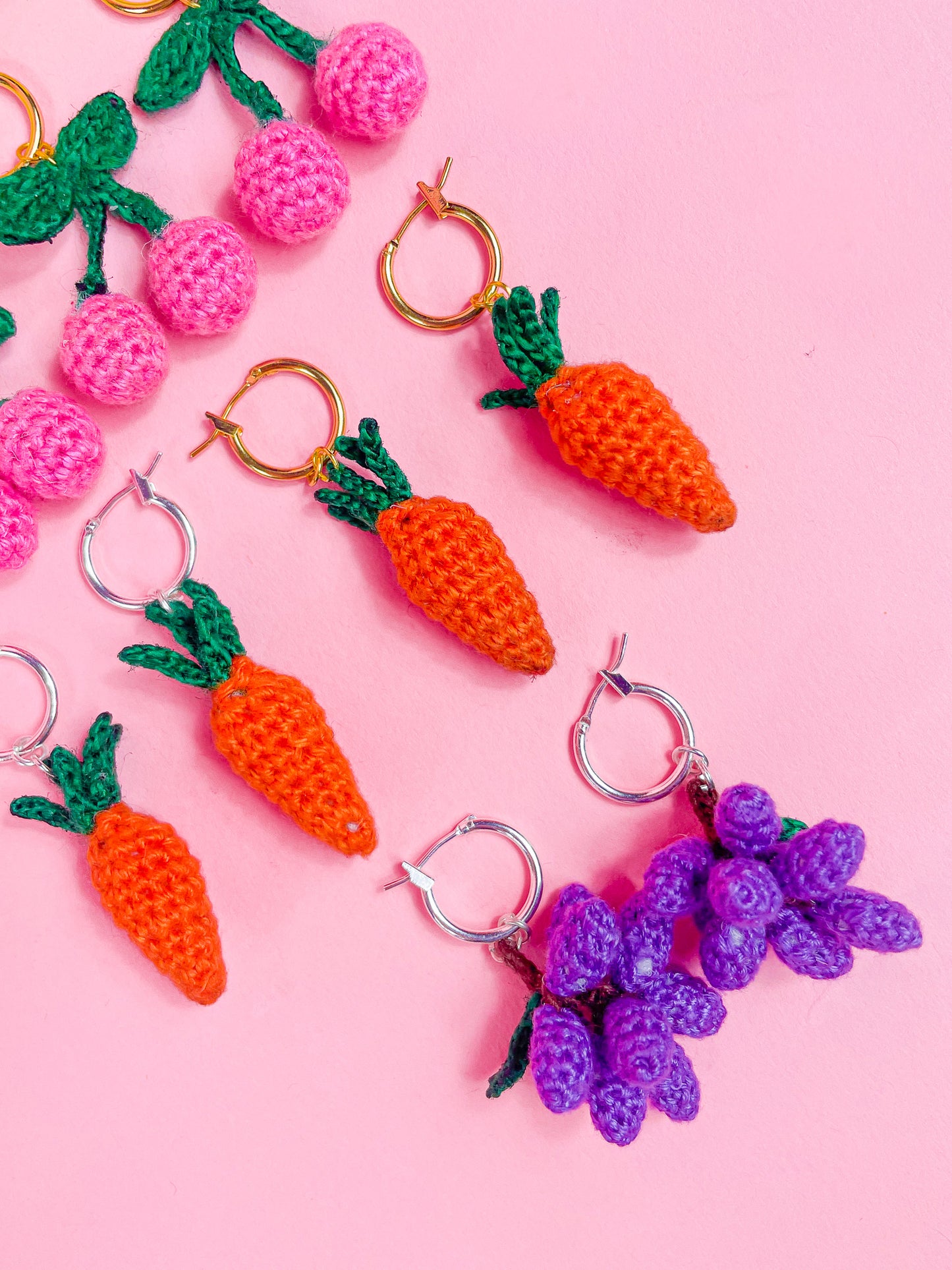 Five pairs of crocheted earrings in different fruit shapes (cherries, carrots and grapes), all lying together in rows on a pink background.