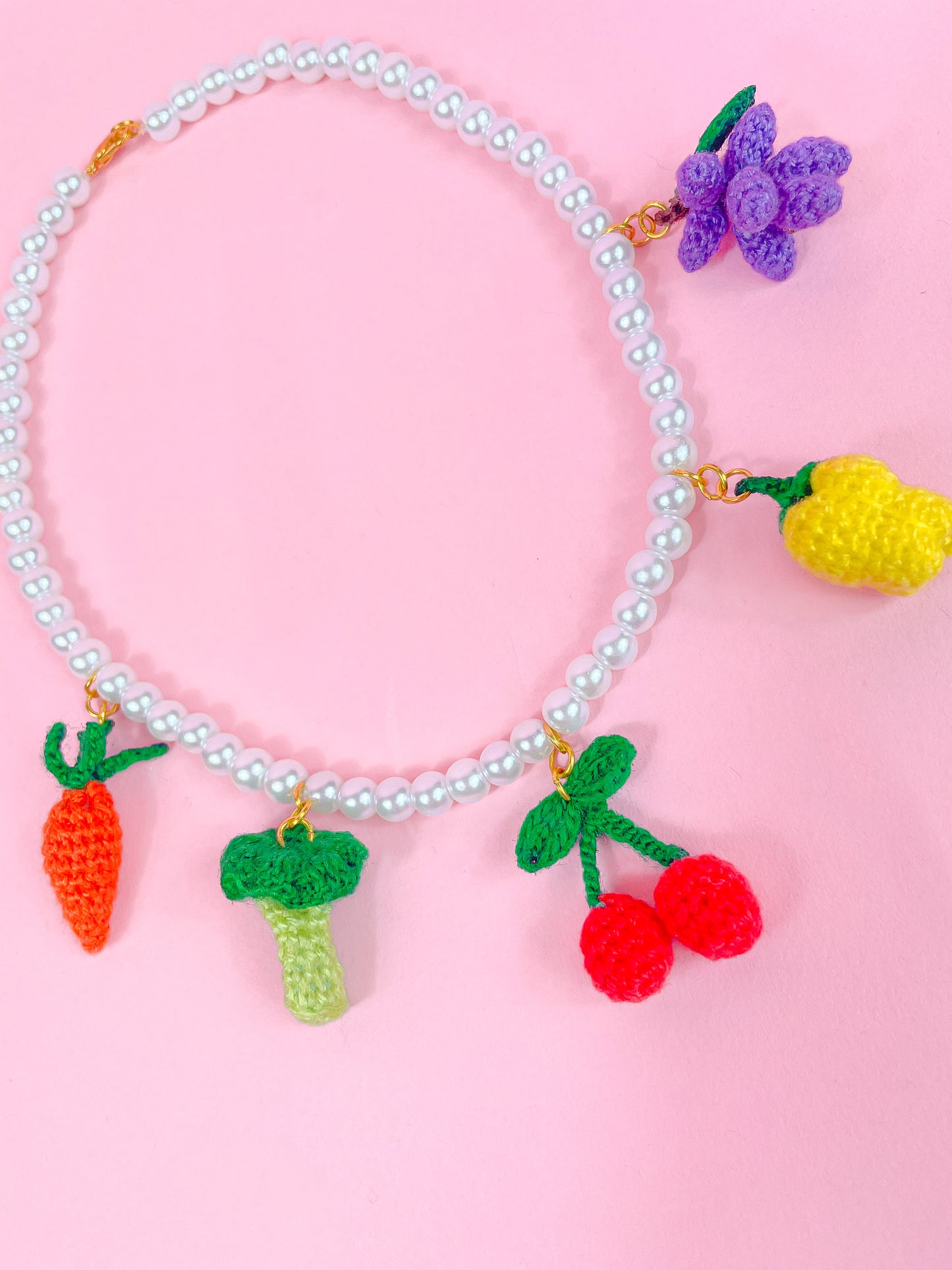 Flat lay side view of a fruit and vegetable crochet beaded choker with a carrot, broccoli, cherry, bell pepper and grape attached around a pearl beaded necklace.