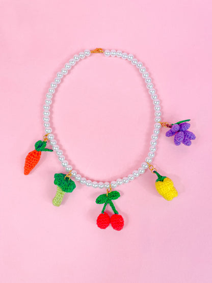 Flay lay of a pearl beaded necklace with a gold lobster clasp and metal rings attaching crochet fruit and vegetable shaped beads to it.