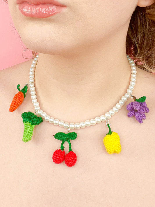 Close up of a woman wearing a pearl beaded necklace with micro crochet fruit and vegetable shaped beads attached to it (carrot, broccoli, cherry, yellow bell pepper, purple grapevine).