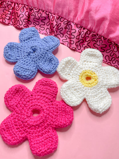 Three crochet scrunchies shaped like daisies in pink, blue and white lying together on a pink background.