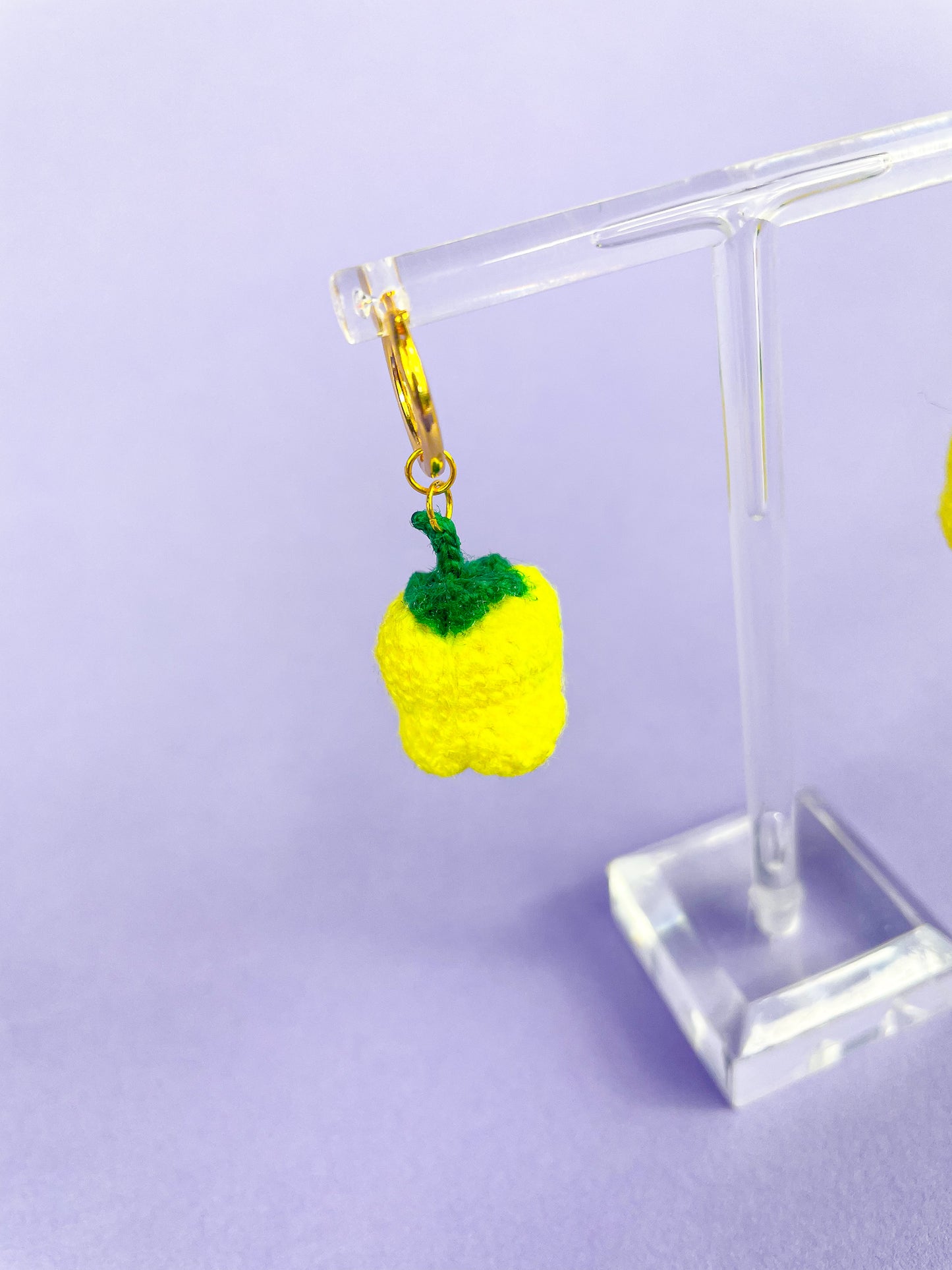 Close view of one small gold hoop earring with a crochet yellow bell pepper hanging from it in front of a purple background.