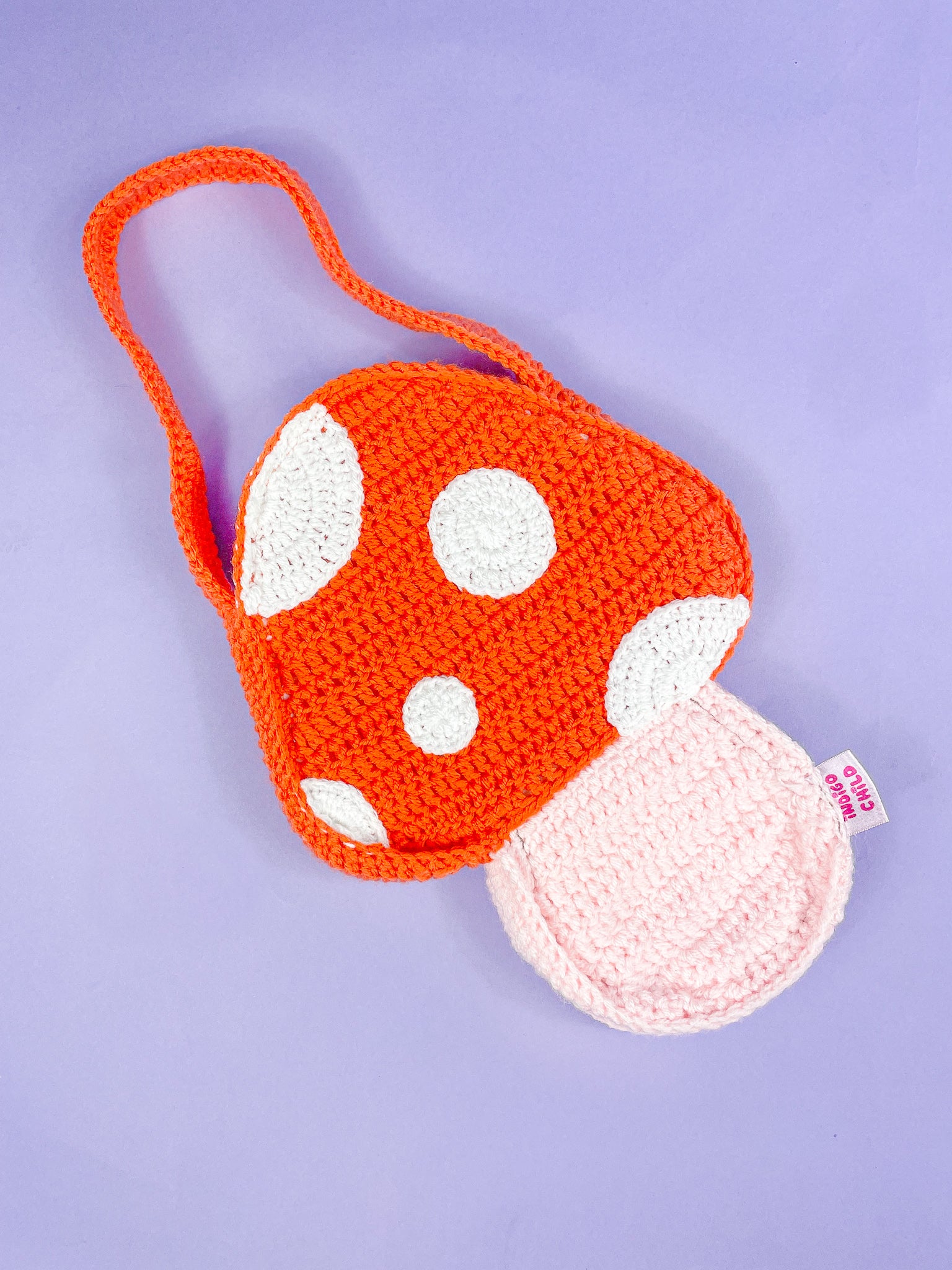 A mushroom shaped bag in orange with crochet dots sewn on lying flat on a lilac background.