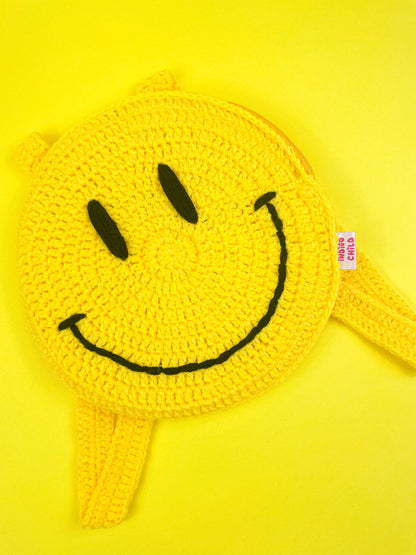 Close view of a yellow circle backpack with a hand stitched smiley face on the front lying on a yellow background.