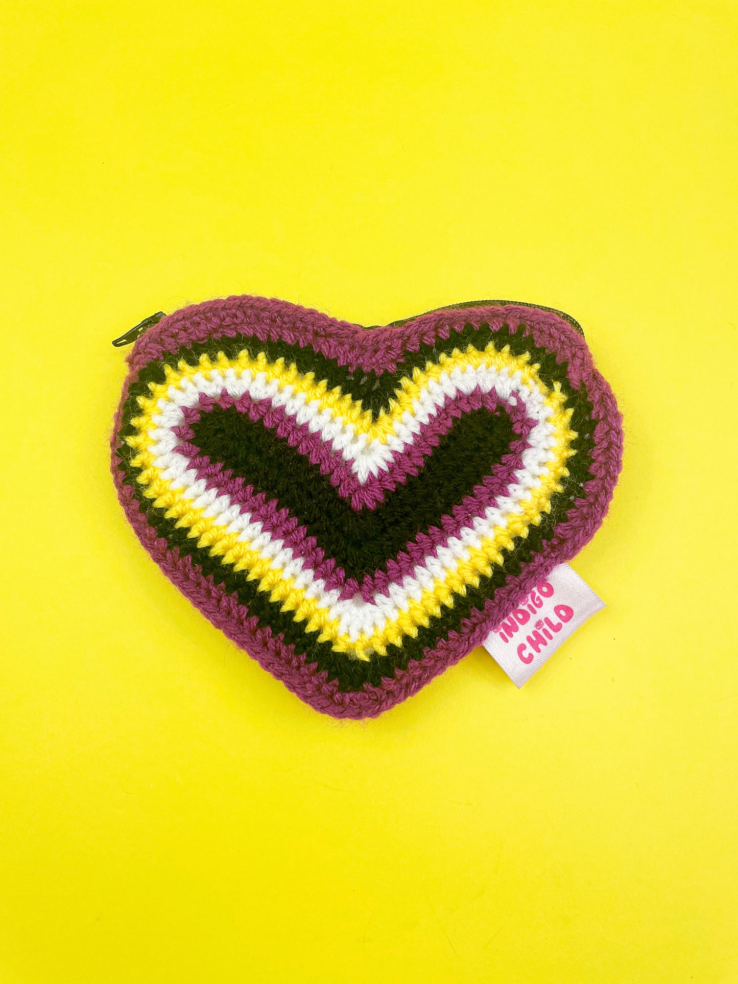 A crochet purse shaped like a heart with a rainbow of non binary flag colours (black, purple, white and yellow).