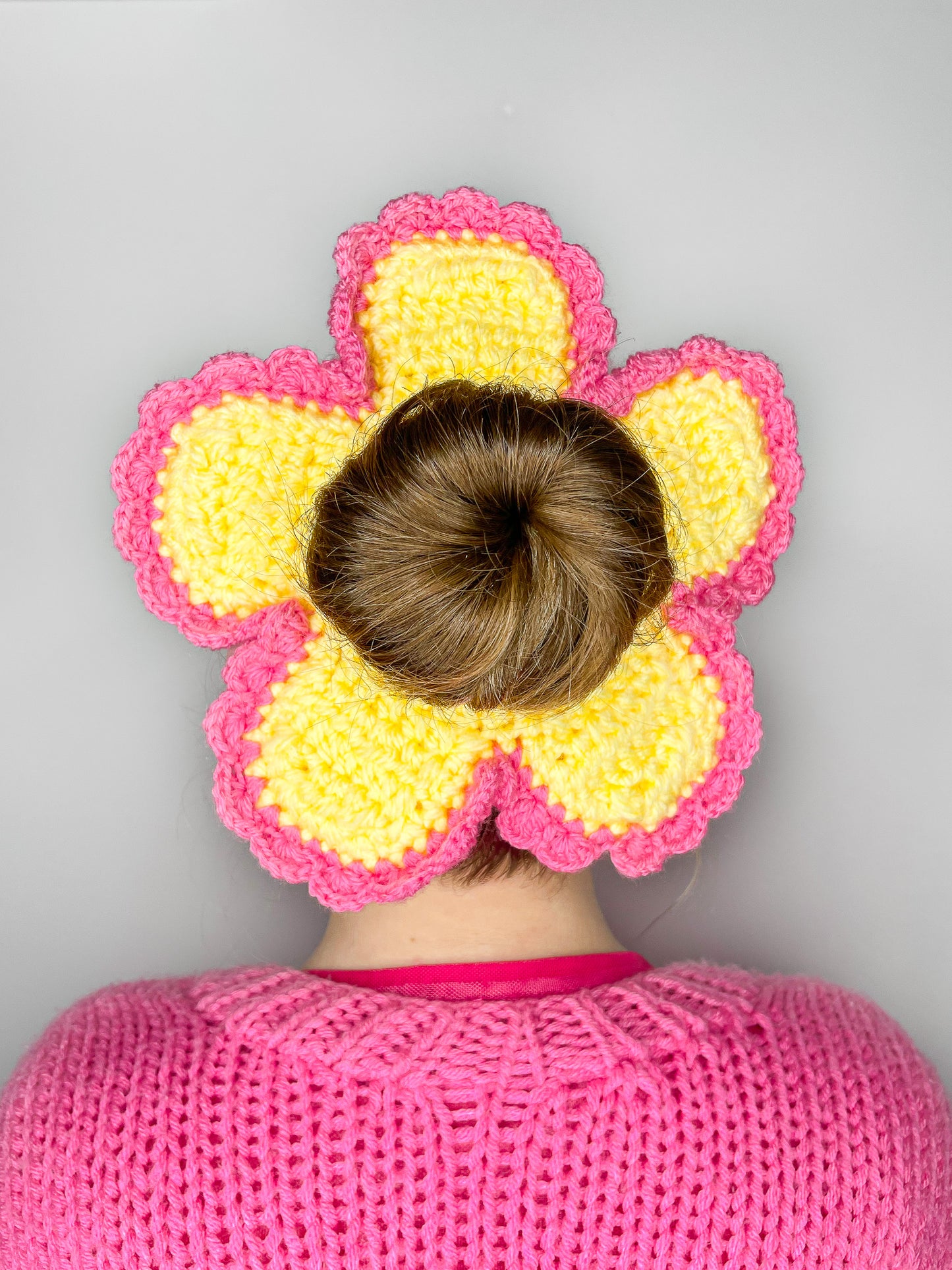 Back view of a woman wearing a daisy shaped crochet scrunchie in yellow with pink frills round the edge, paired with a pink knitted cardigan.