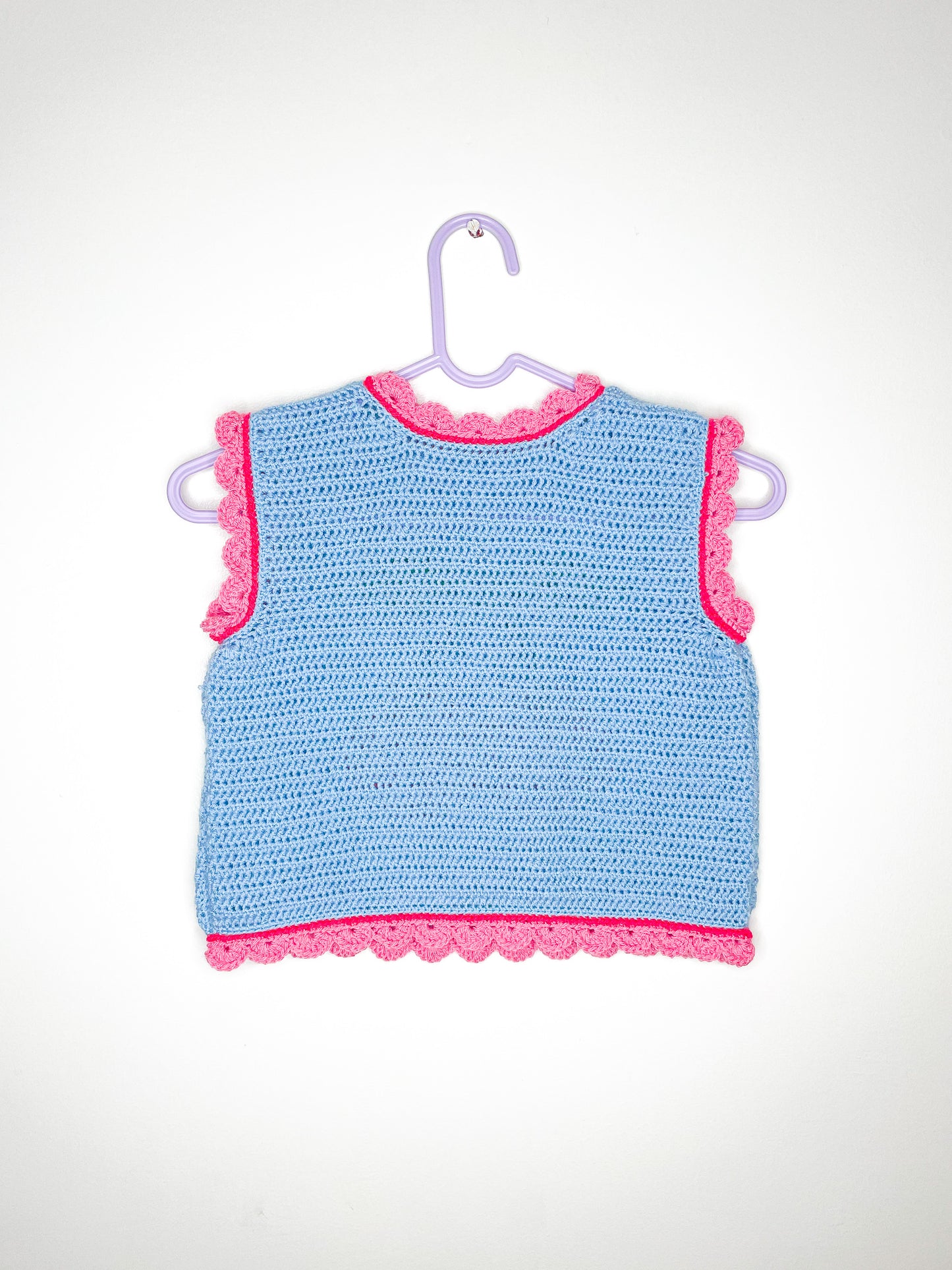 Back view of a vest top in baby blue with pink frilly edges hung on a lilac hanger against a white wall.