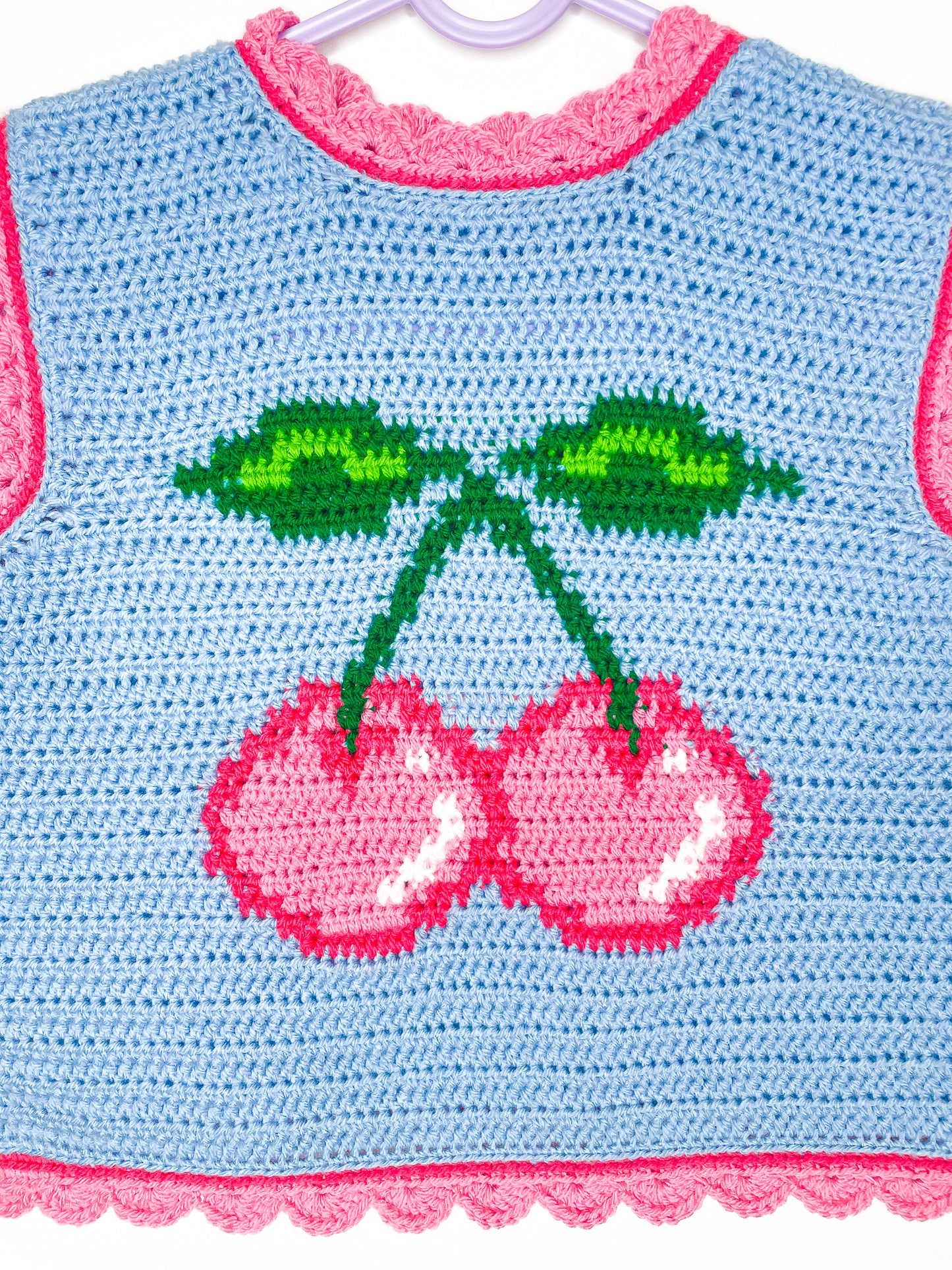 Close view of a tapestry crochet vest in baby blue with a pink cherry on the front and pink edges.