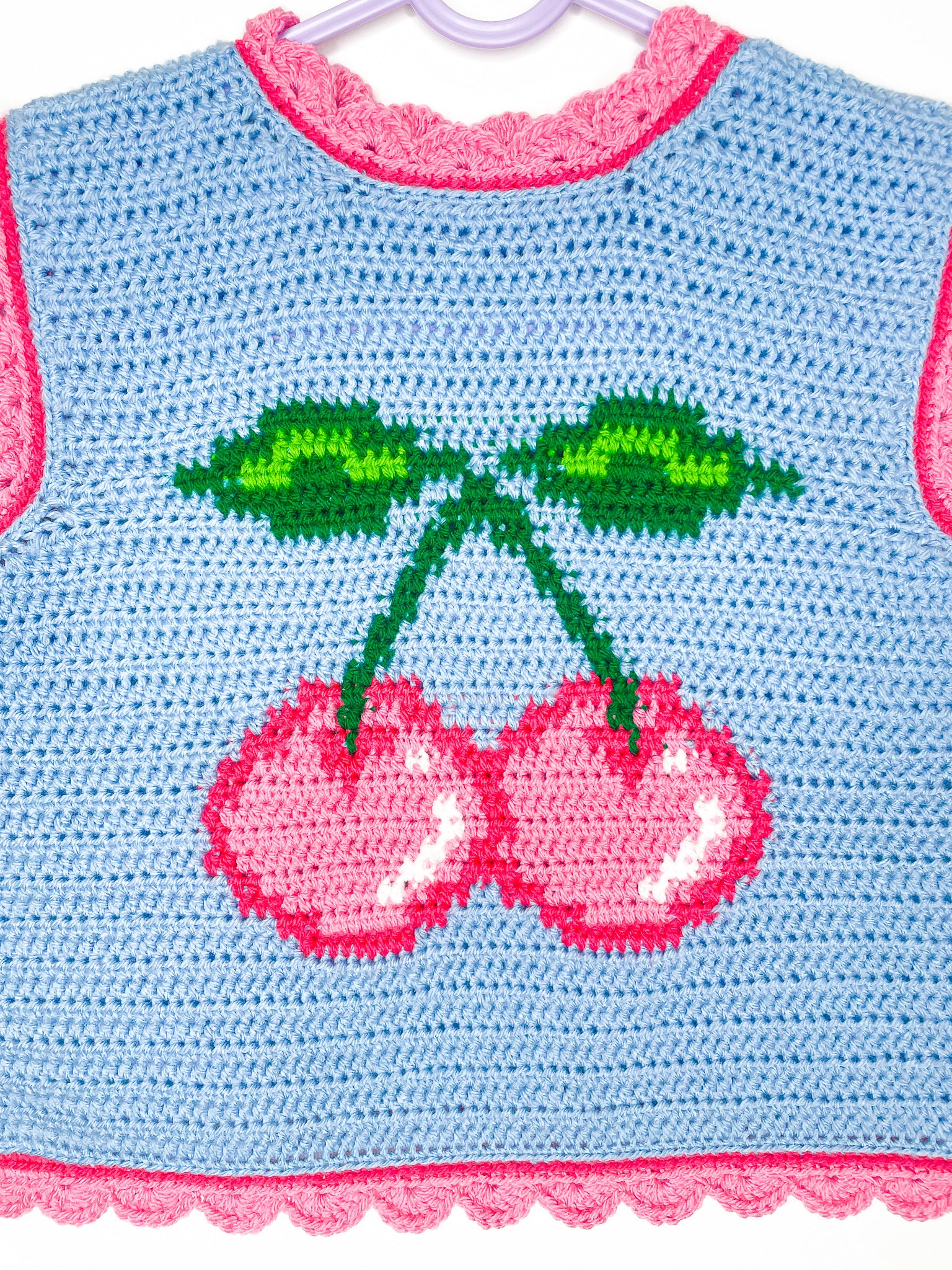 Close view of a tapestry crochet vest in baby blue with a pink cherry on the front and pink edges.