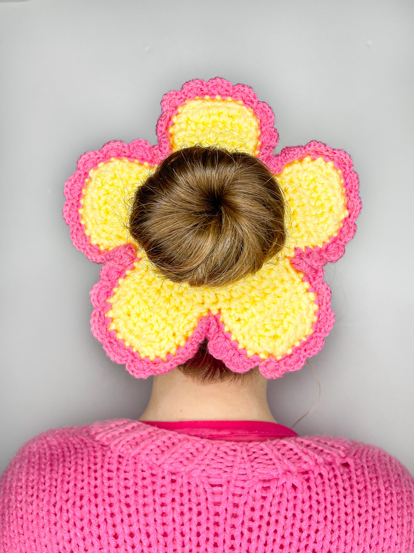 Close view of a woman wearing a daisy shaped scrunchie in yellow with pink frilly edges and a matching pink cardigan.