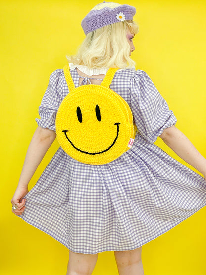 Back view of a woman posing wearing a circle shaped backpack with a happy face on it, paired with a lilac gingham dress.