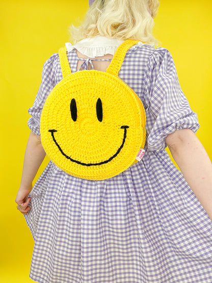 Back view of a woman wearing a yellow crochet backpack shaped like a circle with a smiley face on the base.