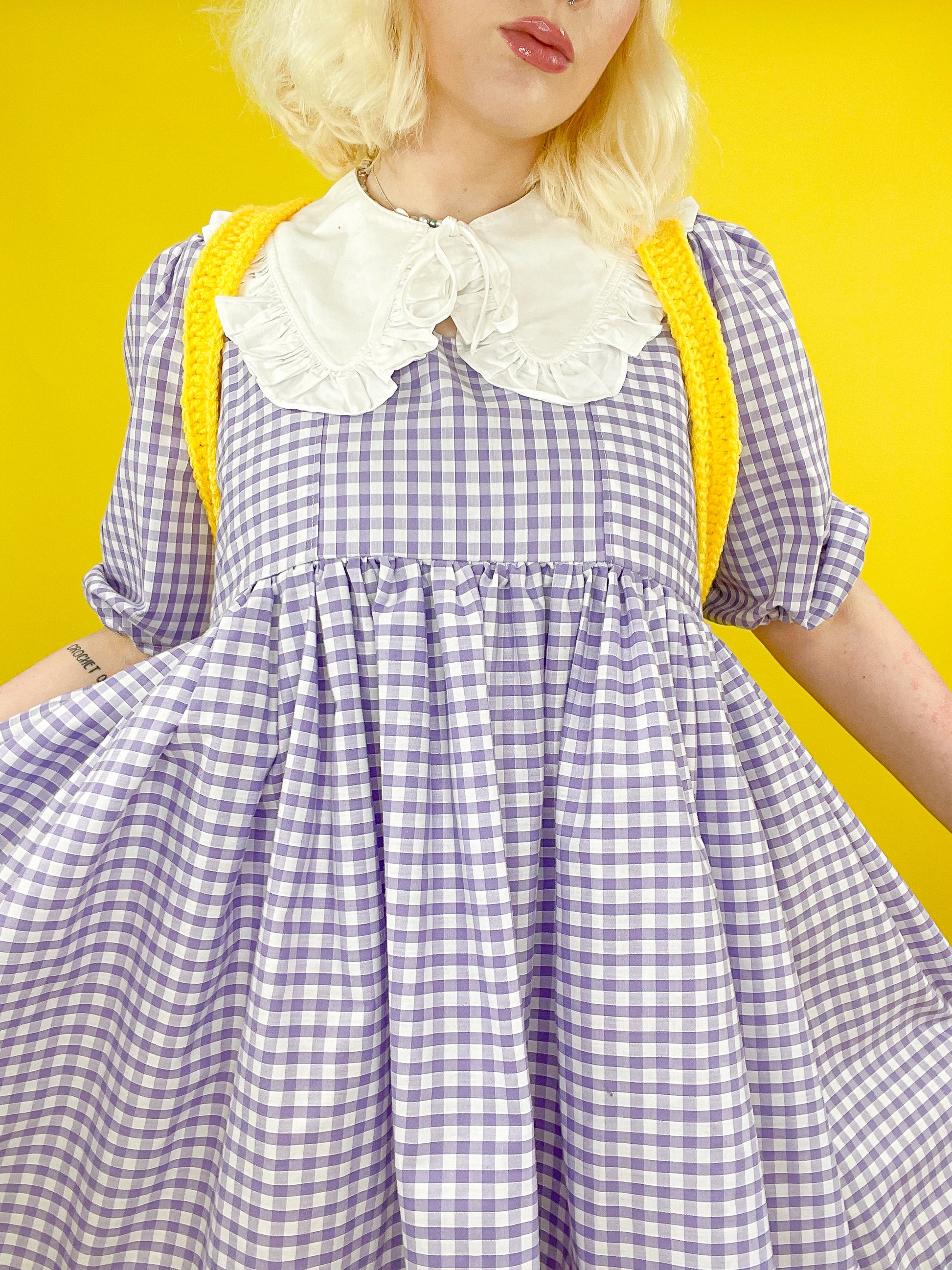 Front view of a woman wearing a yellow backpack on her shoulders with a gingham purple puffy dress.