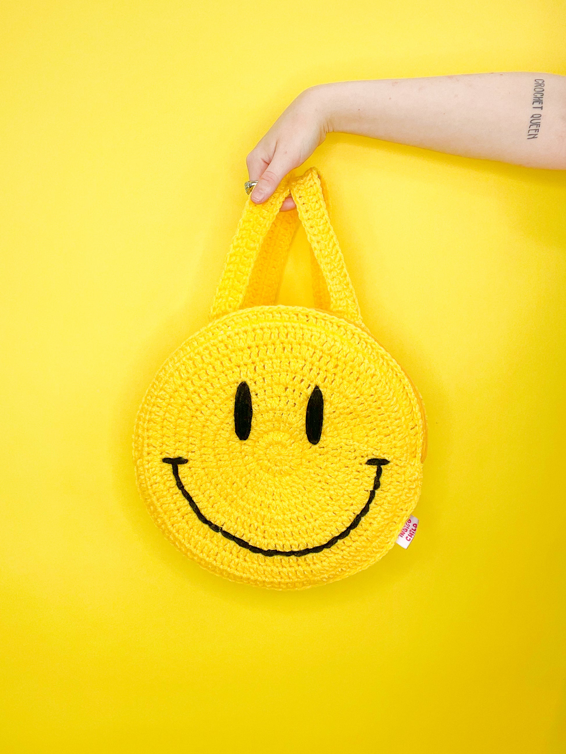 A hand holding a smiley crochet back pack in yellow held in front of a yellow background.