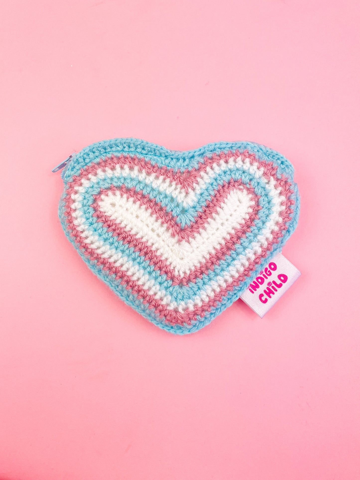 A heart shaped purse with resembling the transgender flag (white, pink and blue) in a rainbow, lying on a pink background.