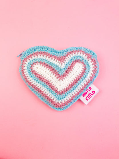 A heart shaped purse with resembling the transgender flag (white, pink and blue) in a rainbow, lying on a pink background.