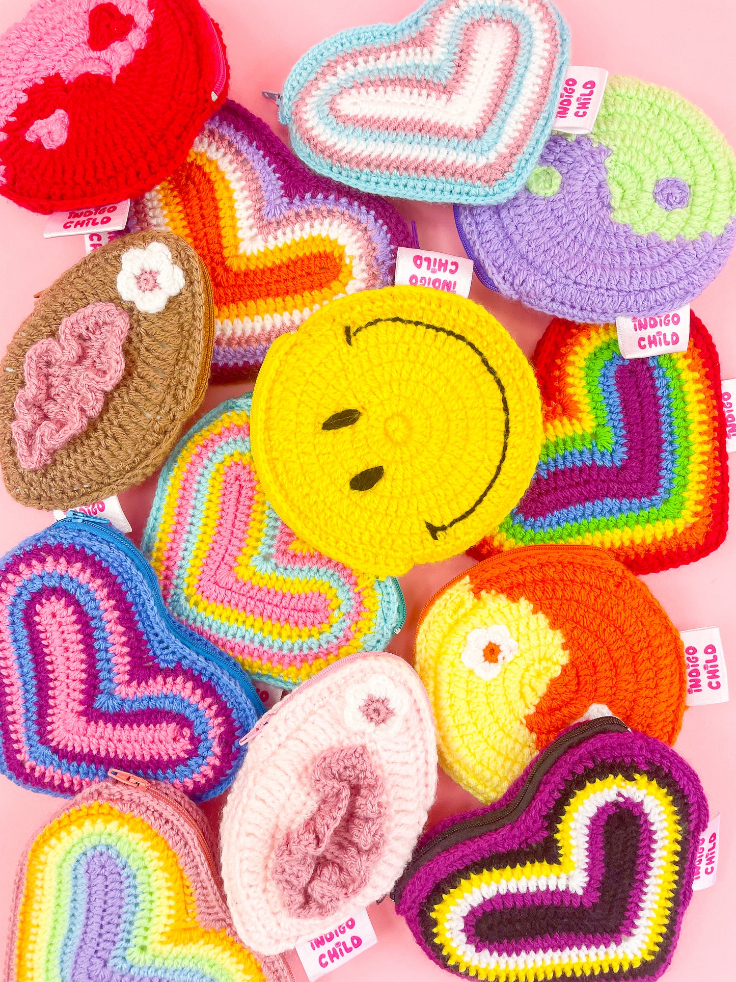 Multiple crocheted coin purses shaped like hearts, smiley faces, yin yangs and vulvas, lying together on a pink background.