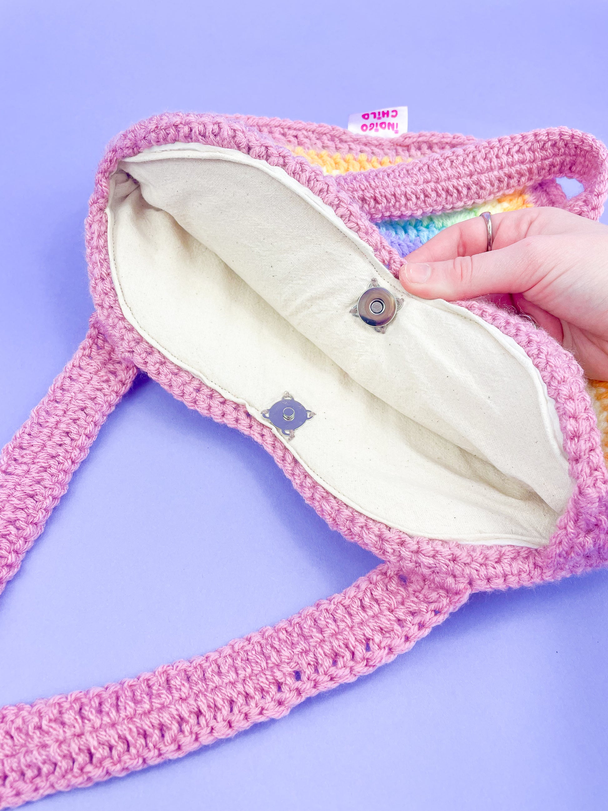 A hand holding one side of the inside of a crochet bag showing the cotton lining and secure magnetic clasps inside it.