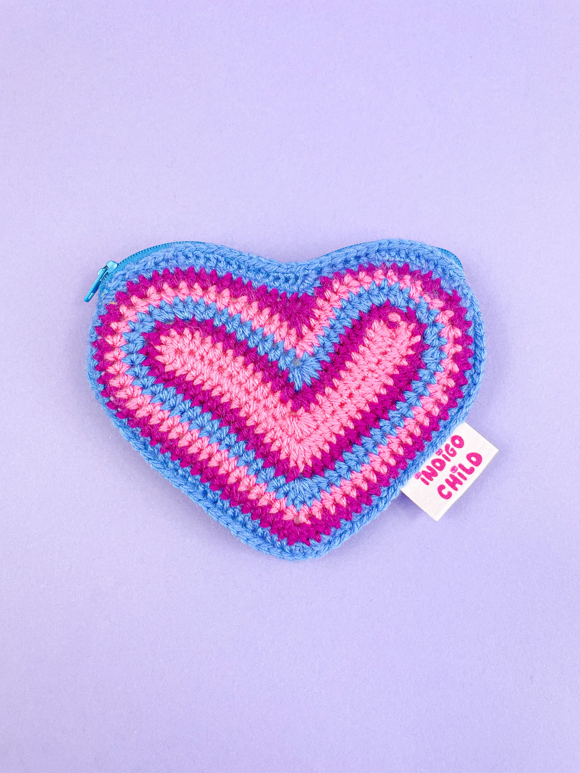 Heart shaped purse in the bisexual flag colours (pink, purple and blue) with a sewn Indigo Child outer tag, lying flat on a purple background.