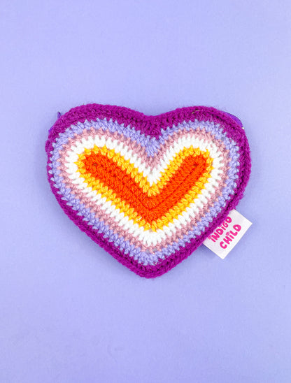 A heart shaped purse with a lesbian flag design lying on a purple background.