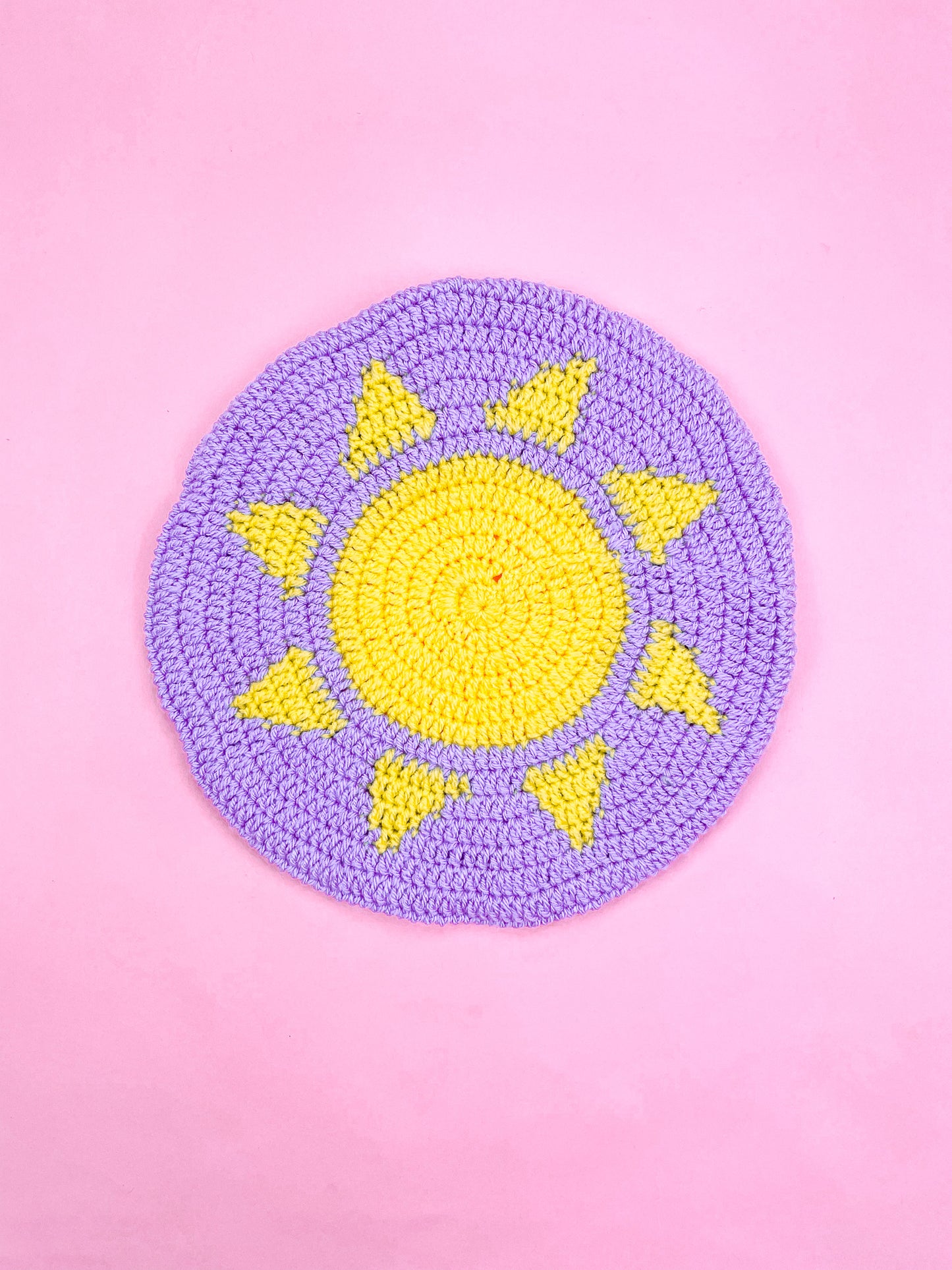 A beret hat in purple with a tapestry crochet yellow sun on the base lying on a pink background.