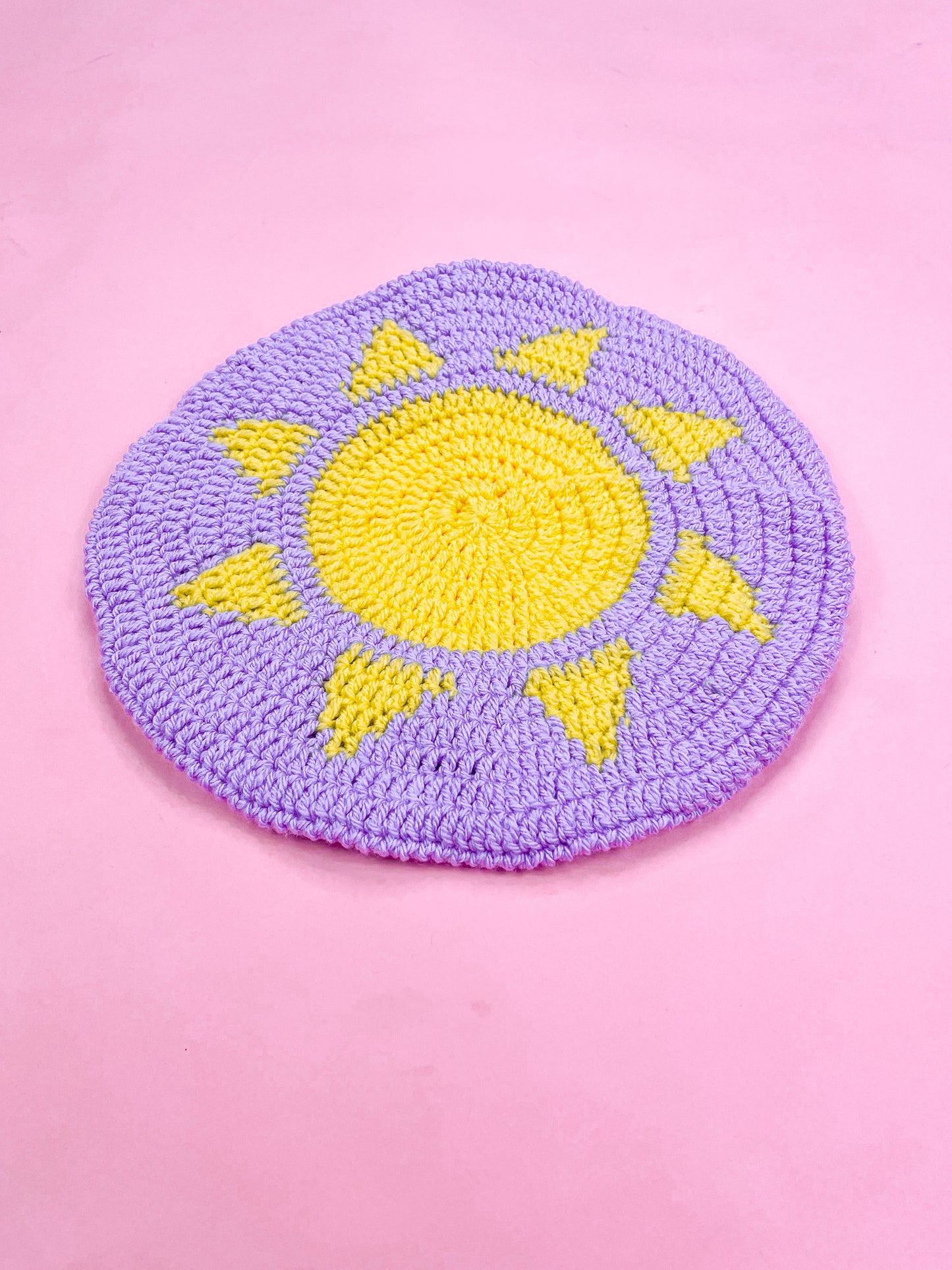 Side view of a purple beret with a sun on the top lying flat on a pink background.