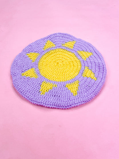 Side view of a purple beret with a sun on the top lying flat on a pink background.