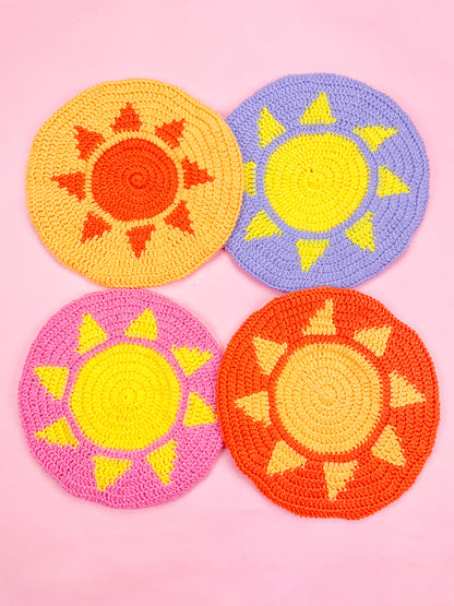 Four crocheted sun berets in yellow, purple, pink and orange lying together on a pink background.