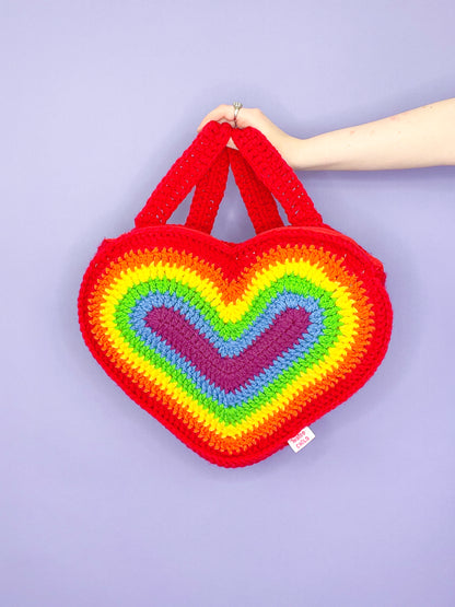 A hand holding a crochet heart backpack in a vibrant rainbow design against a purple background.