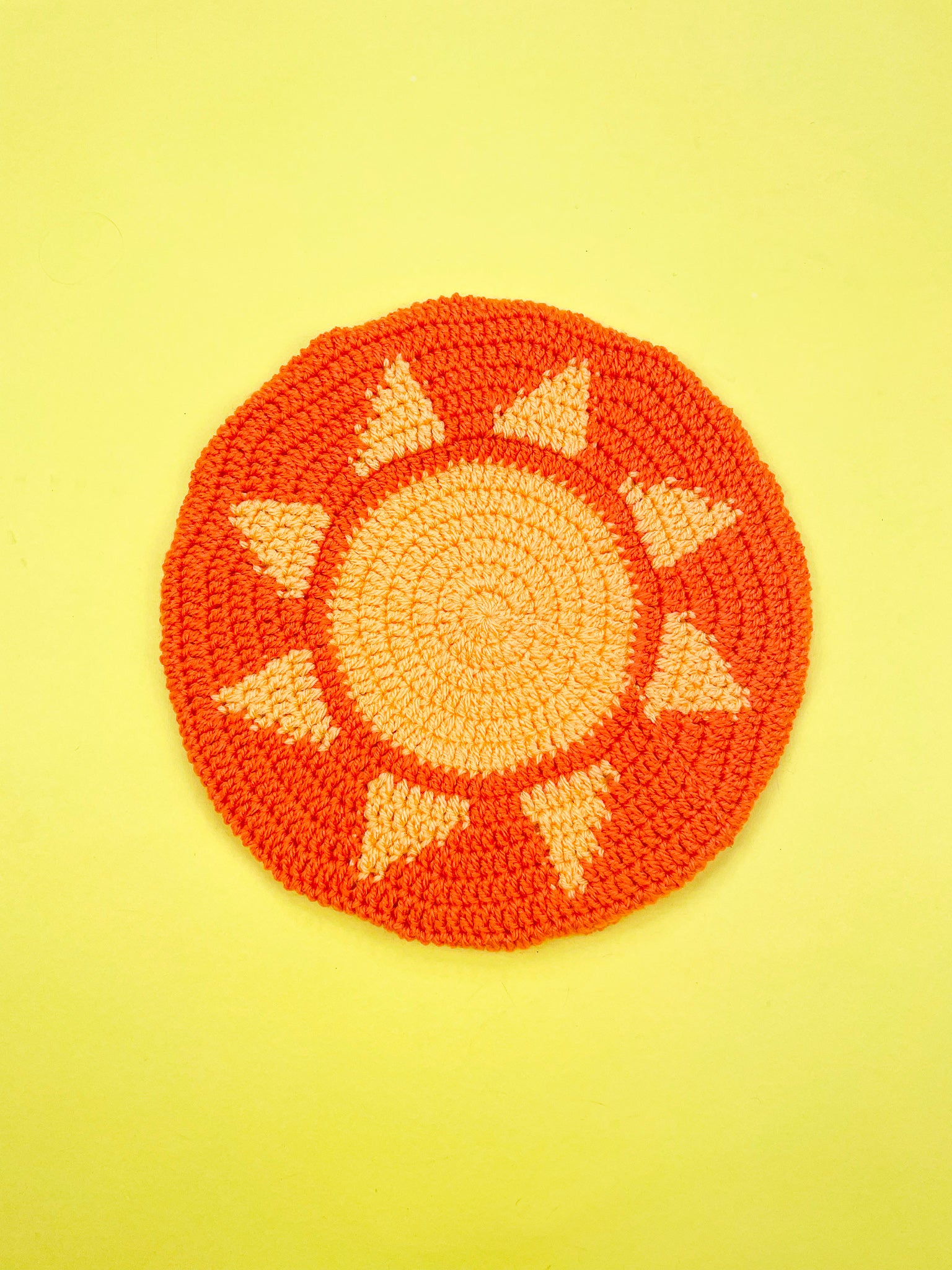 An orange crochet beret hat with a yellow sun in the middle placed on a yellow background.