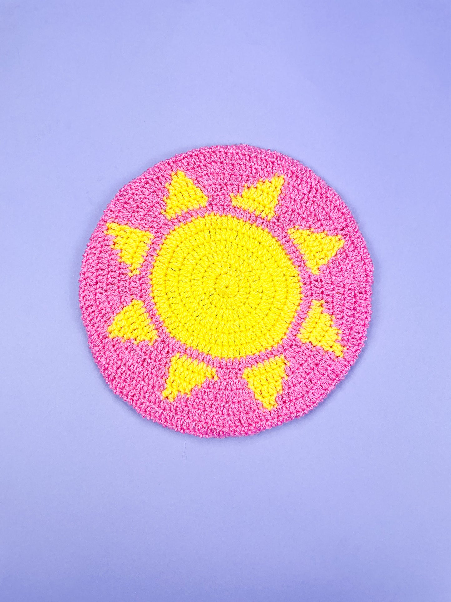 A pink beret with a yellow sun in the centre placed on a purple background.