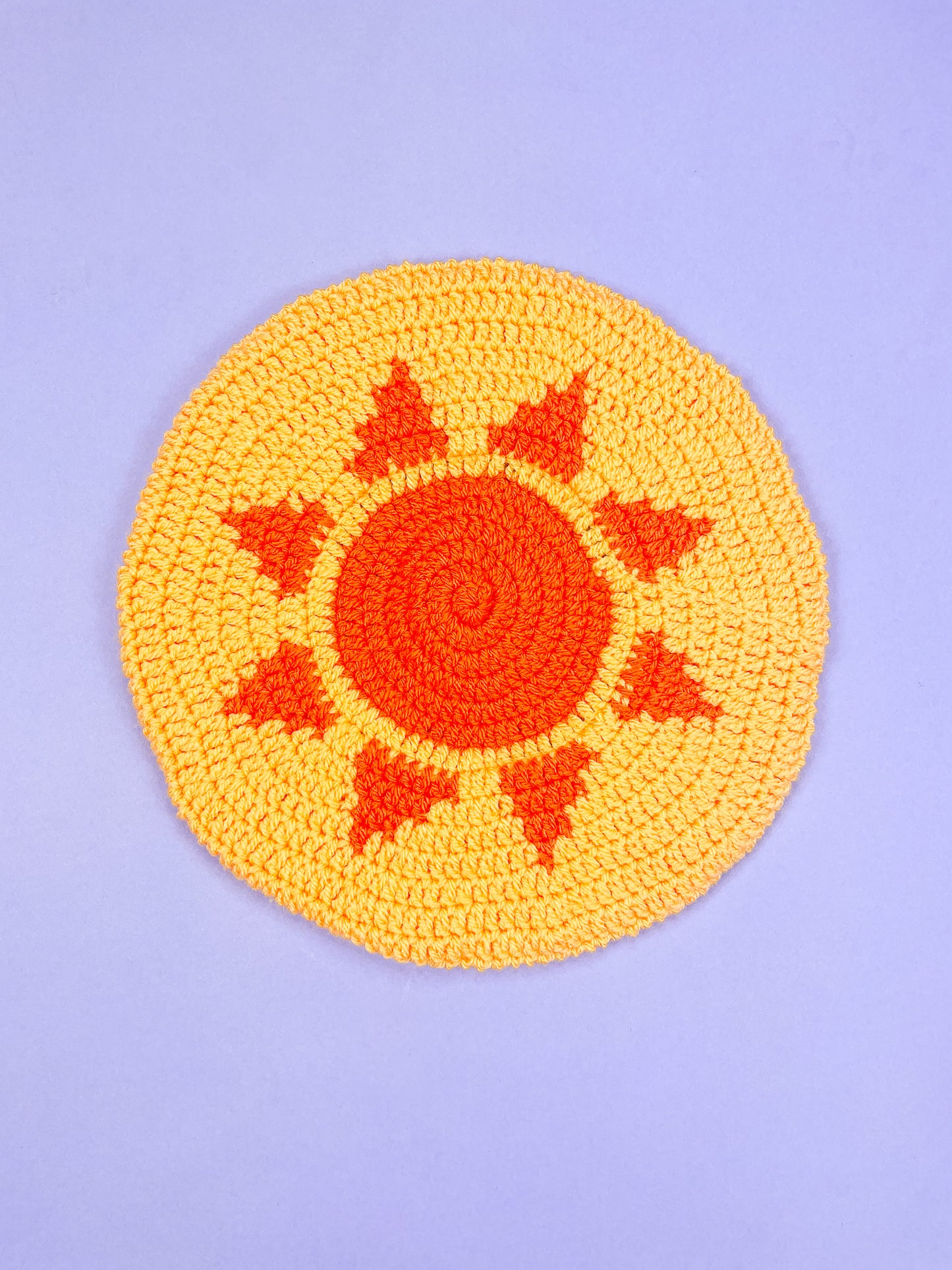 A yellow beret with an orange sun on the base lying on a purple background.