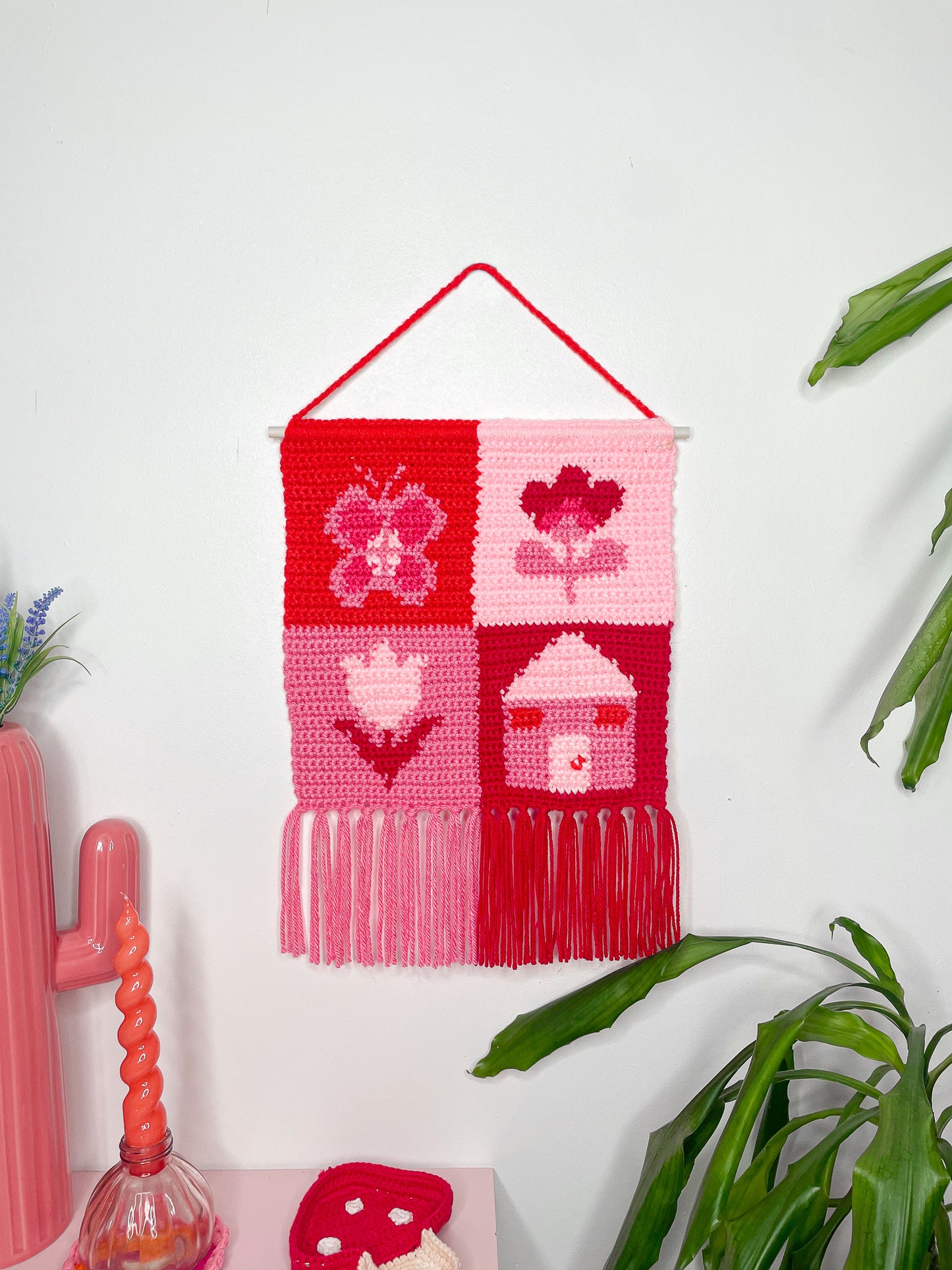 Pink and red tapestry artwork with a patchwork design and tassels attached to the bottom hung on a white wall with a plant, candle, trinket dish and cactus shaped vase next to it on either side.