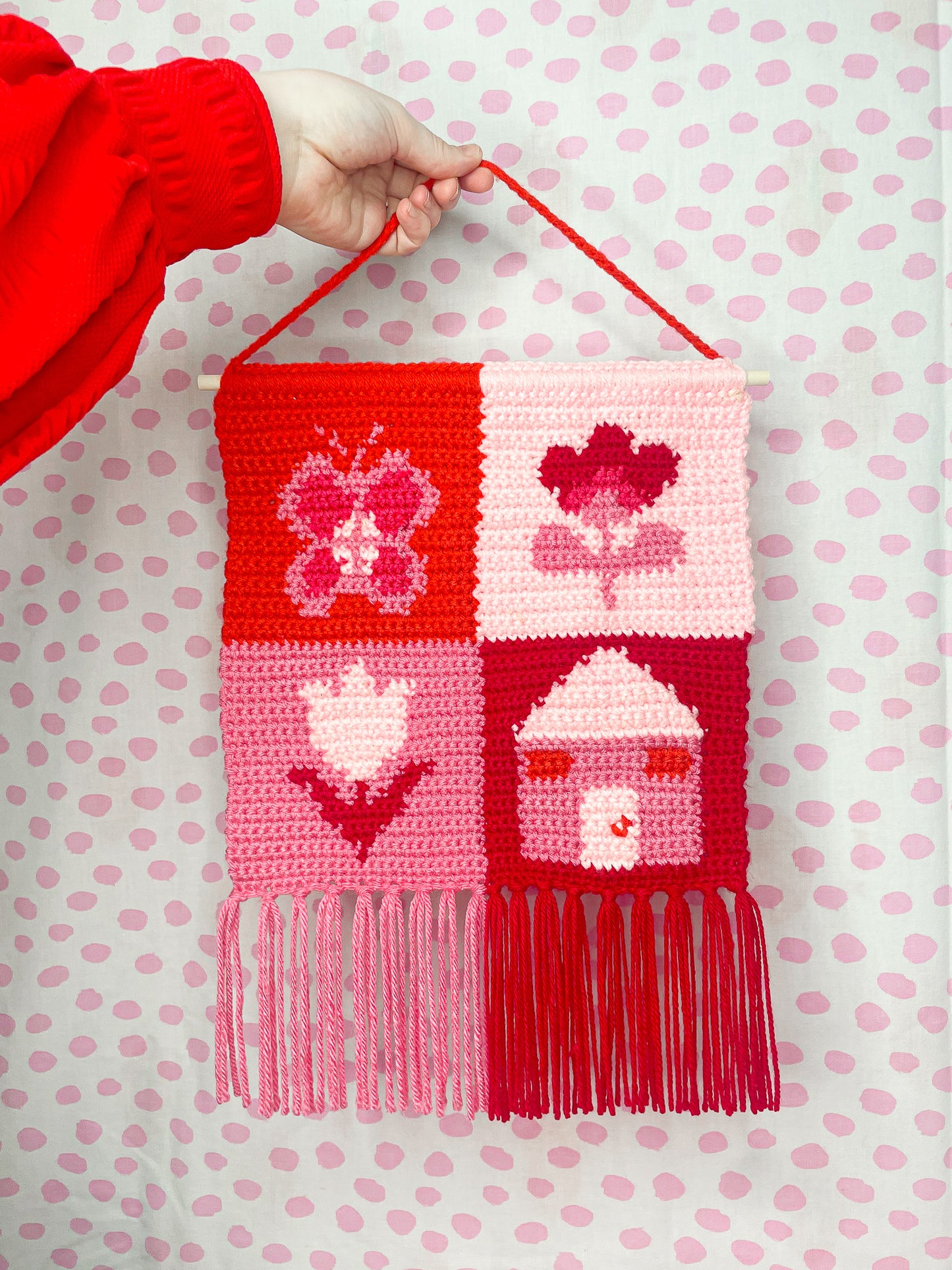 A hand holding a pink and red check wall hanging with a square patchwork design and various images in each square, attached to a wooden rod and decorative tassels added to the bottom edge.