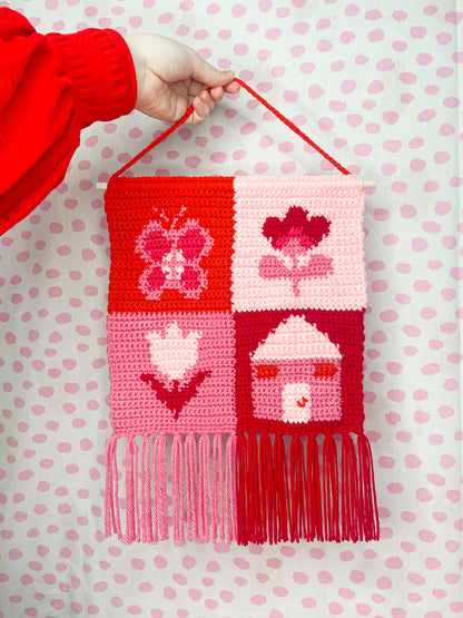 A hand holding a pink and red check wall hanging with a square patchwork design and various images in each square, attached to a wooden rod and decorative tassels added to the bottom edge.