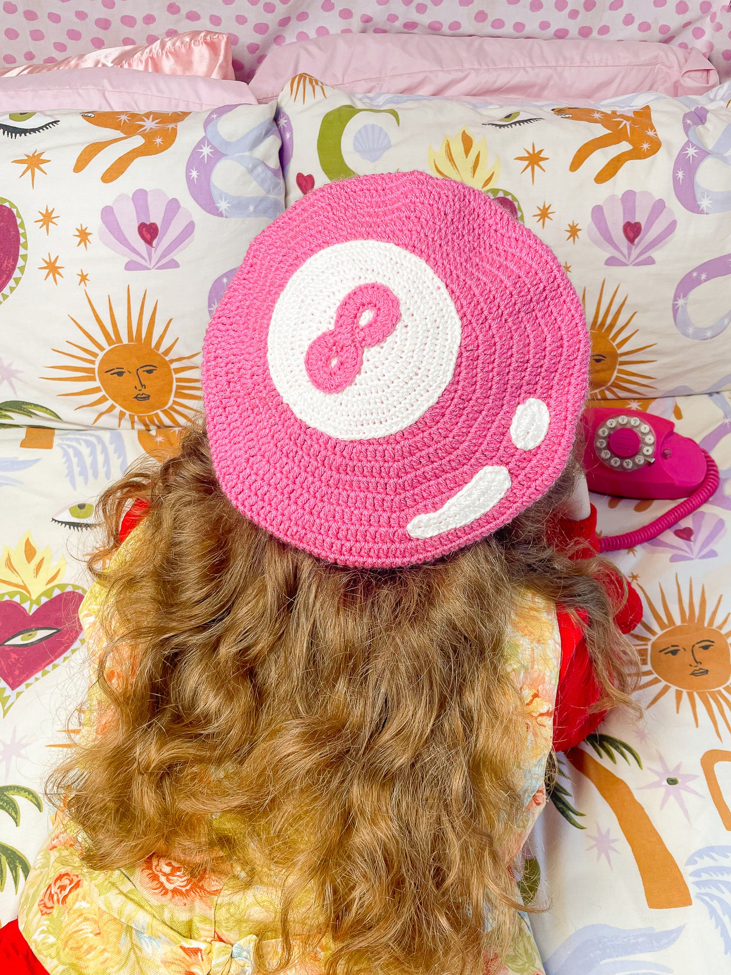 Back view of a woman lying on a bed on her front wearing a pink crochet beret that resembles an 8 ball with a floral waistcoat and red shirt on and funky patterned bed sheets.