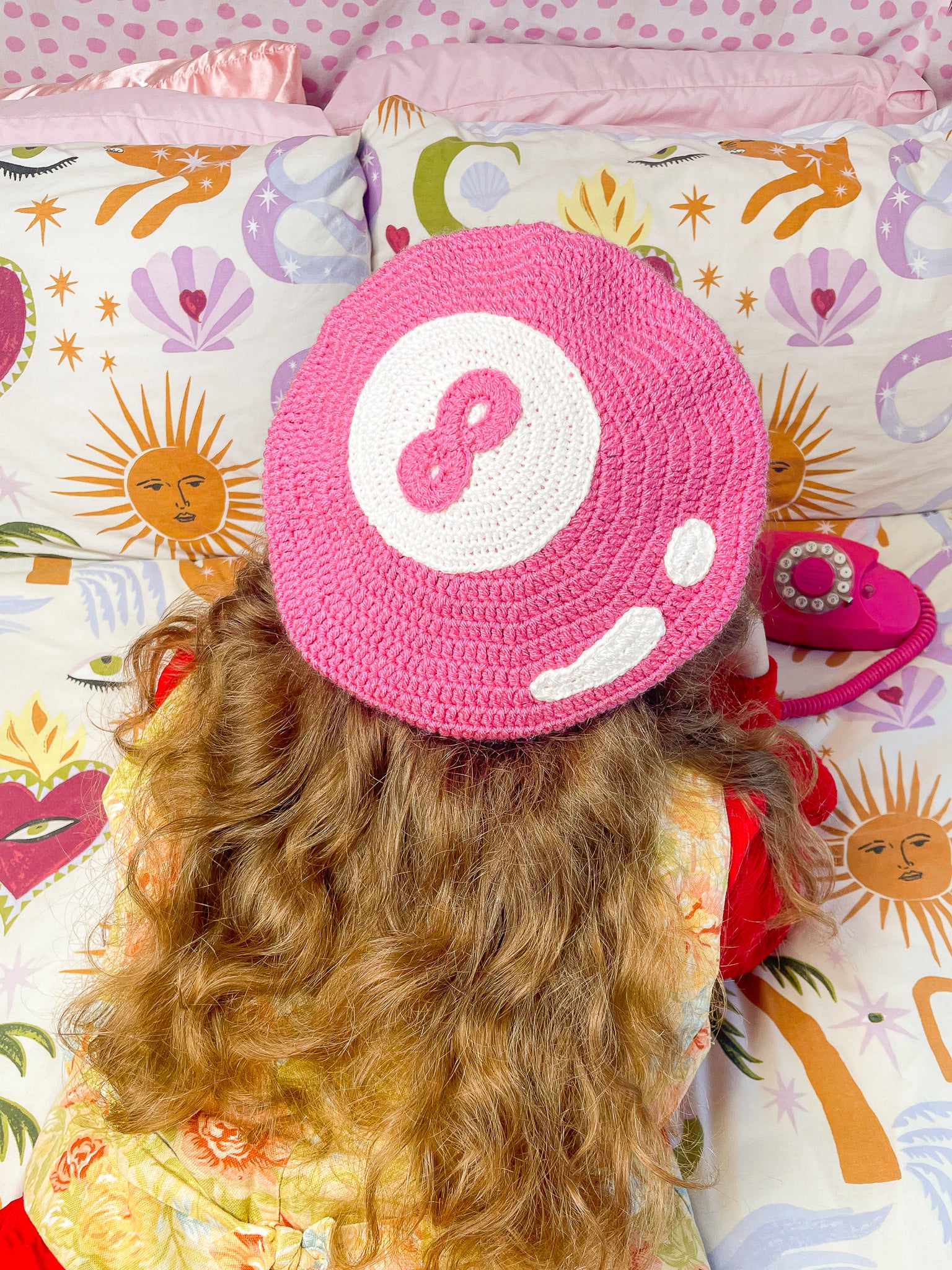 Back view of a woman lying on a bed on her front wearing a pink crochet beret that resembles an 8 ball with a floral waistcoat and red shirt on and funky patterned bed sheets.