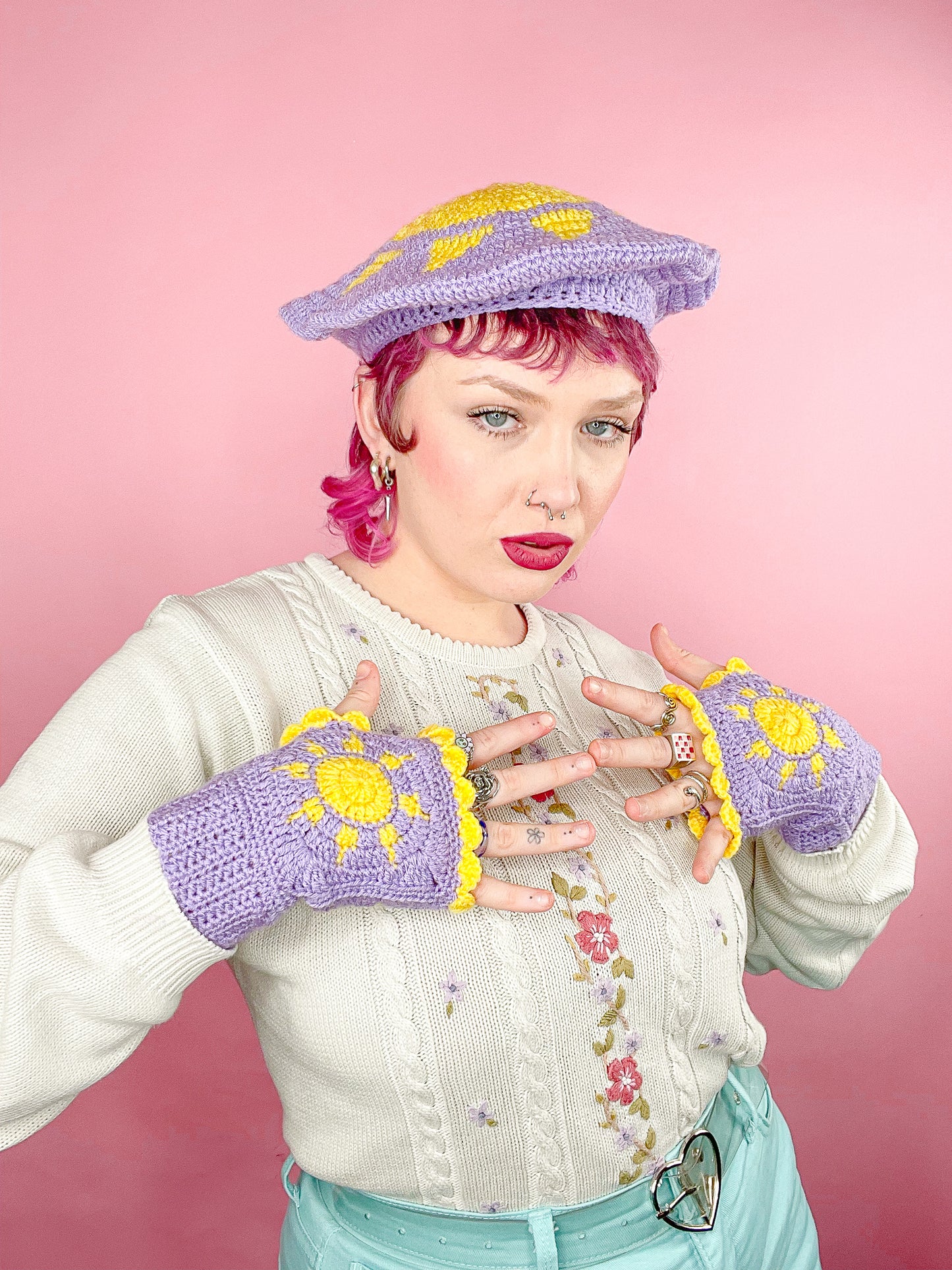 A woman posing wearing a pair of lilac crocheted gloves with suns on them and a matching beret, paired with a granny style jumper.