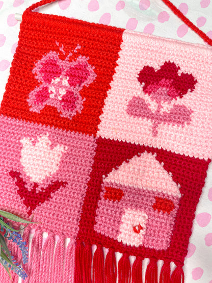 Flay lay of a pink and red check crochet wall hanging with an intricate tapestry design lying on a white and pink dotted sheet.